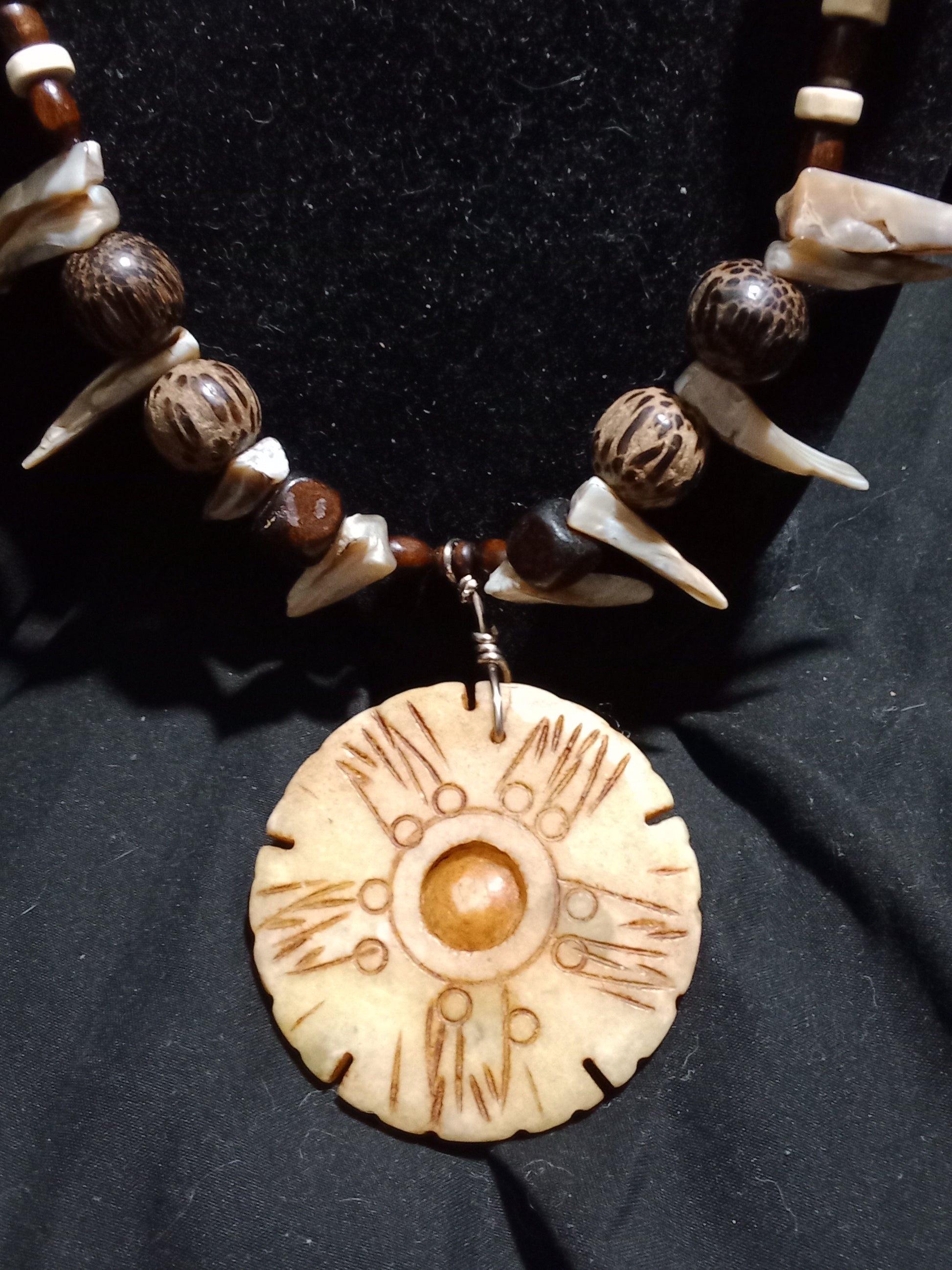 Flex cord beaded necklace with wood beads of various shades, sizes, styles. Accented with natural shell beads in the shape of claws. Bone pendant center piece. Toggle clasp. 23 inches in length.