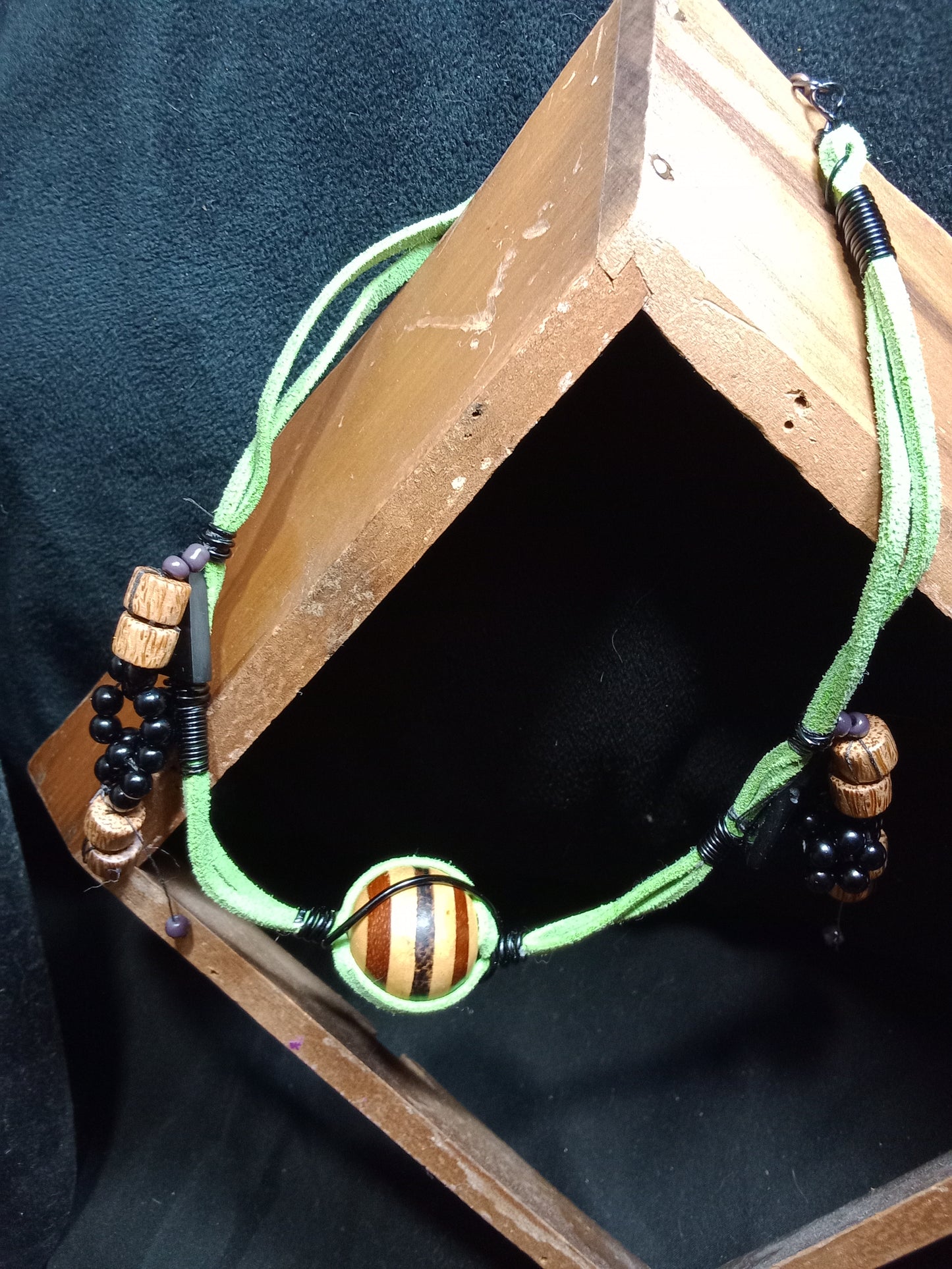 Green suede necklace with wood beads, black round beads, and black triangle beads. One medium sized multicolored striped round bead as center piece. Toggle clasp. 17.5 length